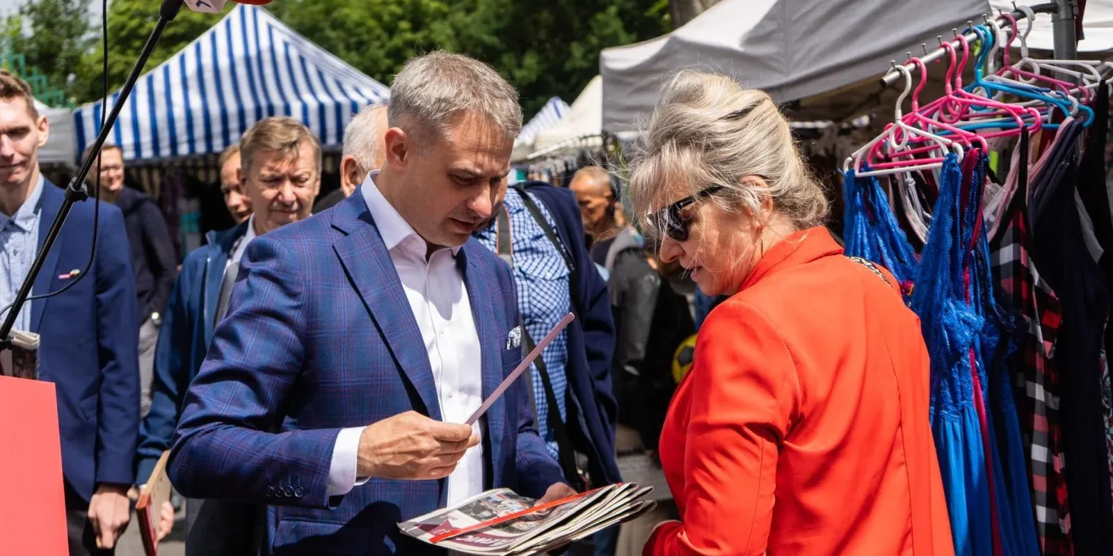 Polityk w granatowo-niebieskim garniturze rozmawia z kobietą w czerwonej marynarce na targu, rozdaje gazety.