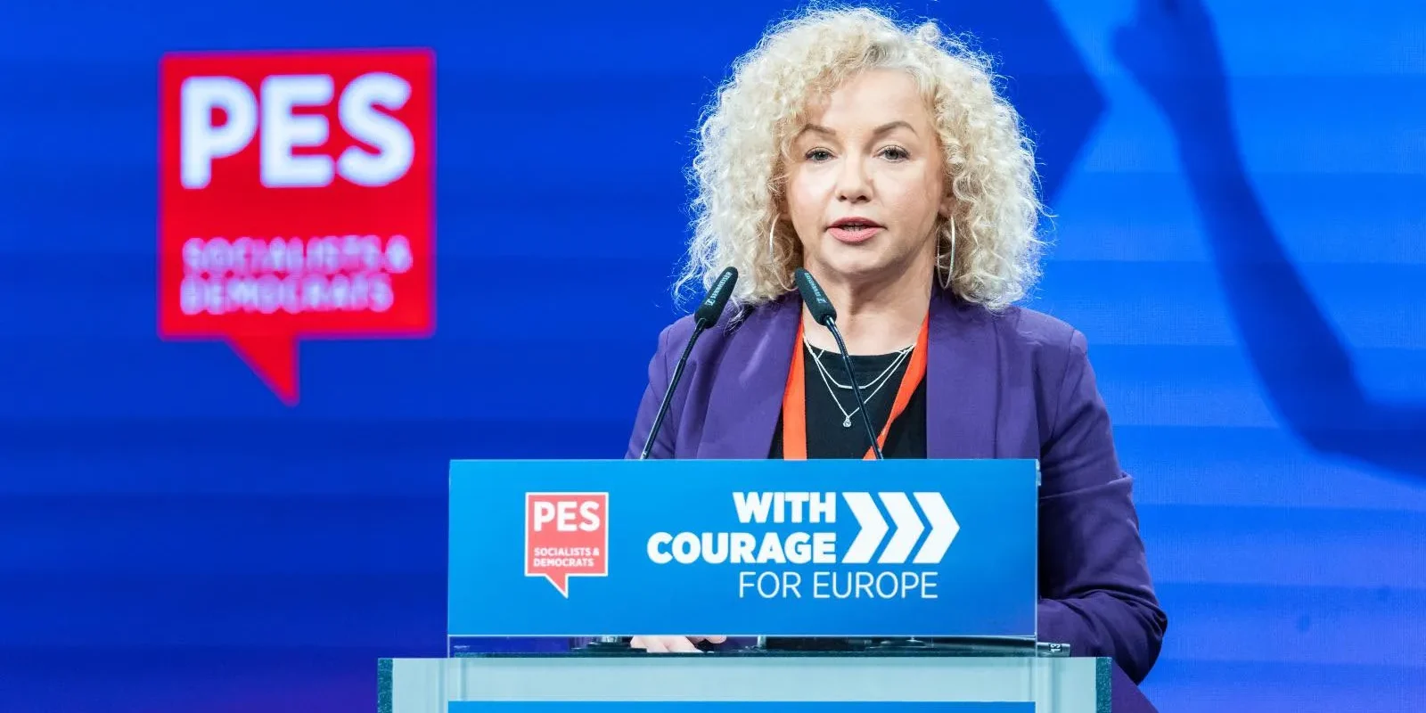 Polityczka z kręconymi blond włosami wygłasza przemówienie z mównicy; w tle widoczne logo PES i napis „With Courage for Europe”.