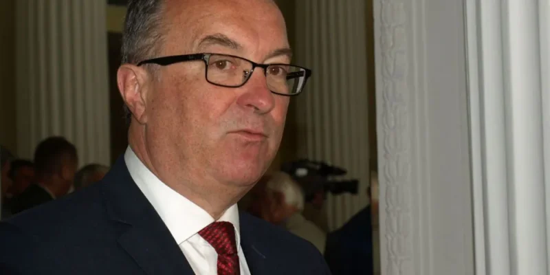 Mężczyzna w okularach, ubrany w ciemny garnitur z czerwonym krawatem, stoi przy ścianie z białymi kolumnami, patrząc w stronę obiektywu.