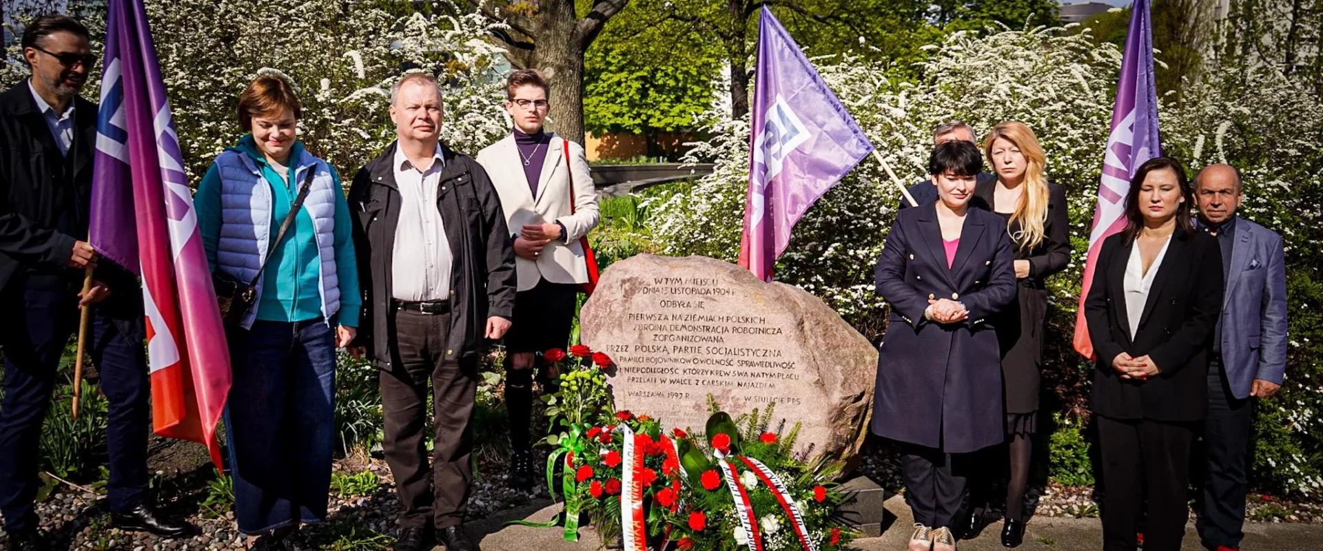 Na zdjęciu widać grupę ludzi podczas uroczystości przy pomniku z kwiatami i flagami, otoczoną zielenią i kwitnącymi krzewami.