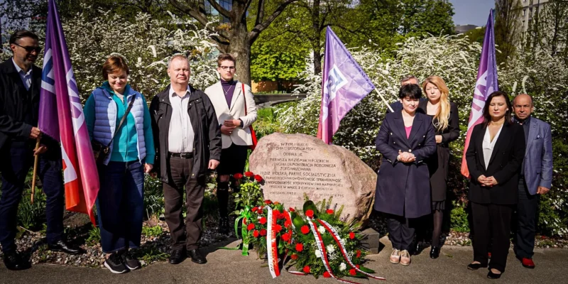 Na zdjęciu widać grupę ludzi podczas uroczystości przy pomniku z kwiatami i flagami, otoczoną zielenią i kwitnącymi krzewami.