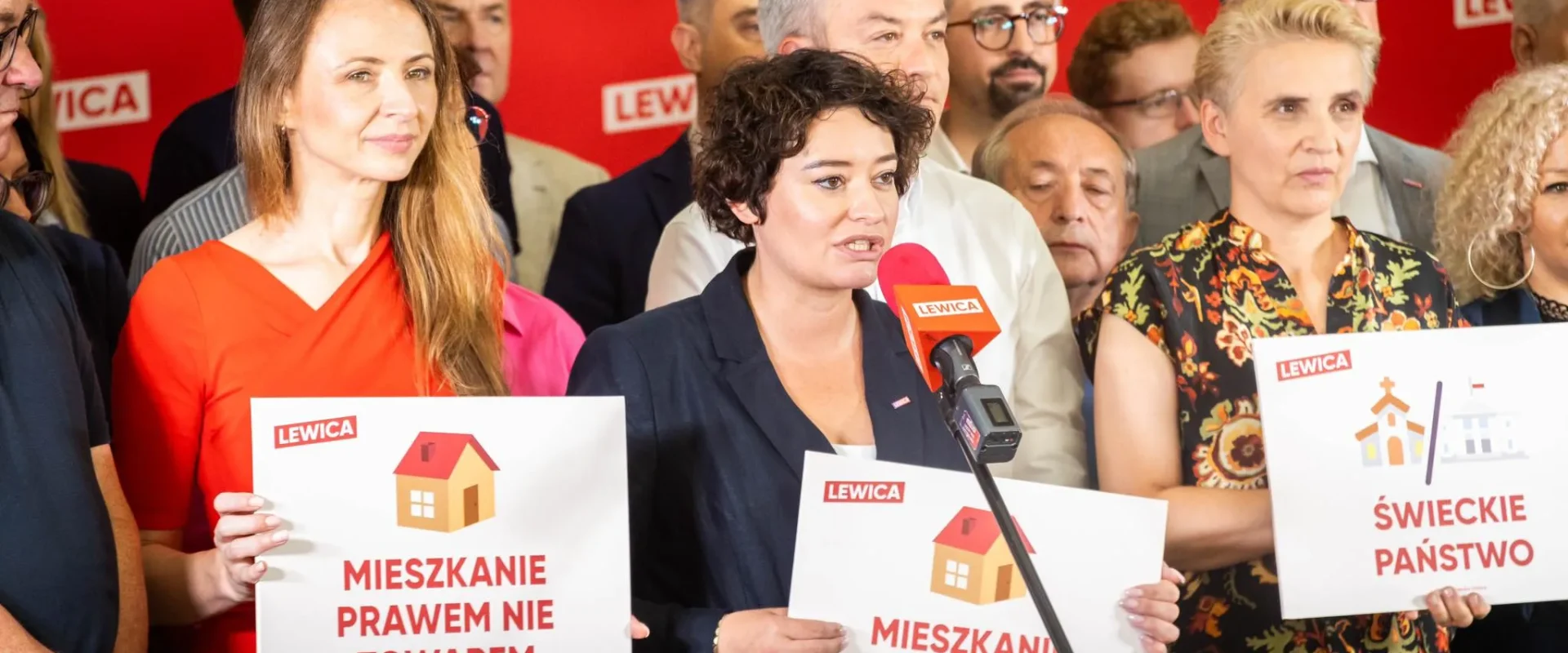 Kobieta przemawia do mikrofonu podczas konferencji Lewicy, otoczona innymi osobami i trzymająca plakat z napisem „MIESZKANIE PRAWEM NIE TOWAREM” na czerwonym tle z logo Lewicy.