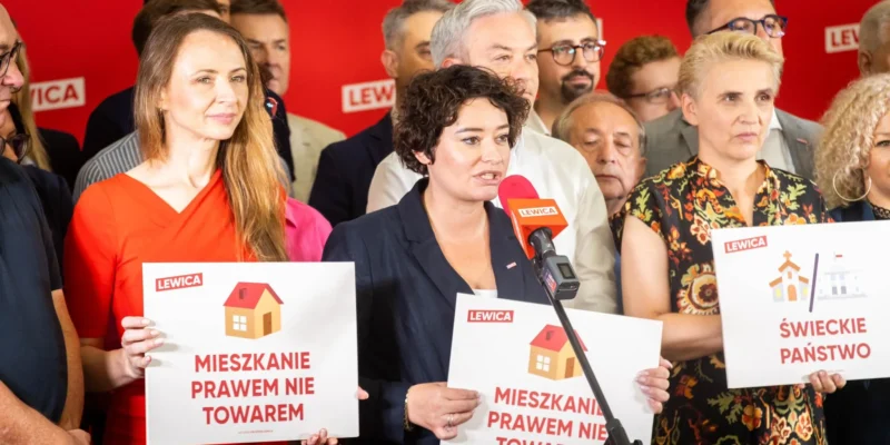 Kobieta przemawia do mikrofonu podczas konferencji Lewicy, otoczona innymi osobami i trzymająca plakat z napisem „MIESZKANIE PRAWEM NIE TOWAREM” na czerwonym tle z logo Lewicy.