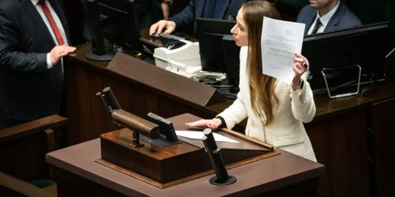 Polityczka w białej marynarce stoi przy mównicy w parlamencie i trzyma kartkę z dokumentem podczas wniosku o wotum nieufności wobec ministra Przemysława Czarnka.