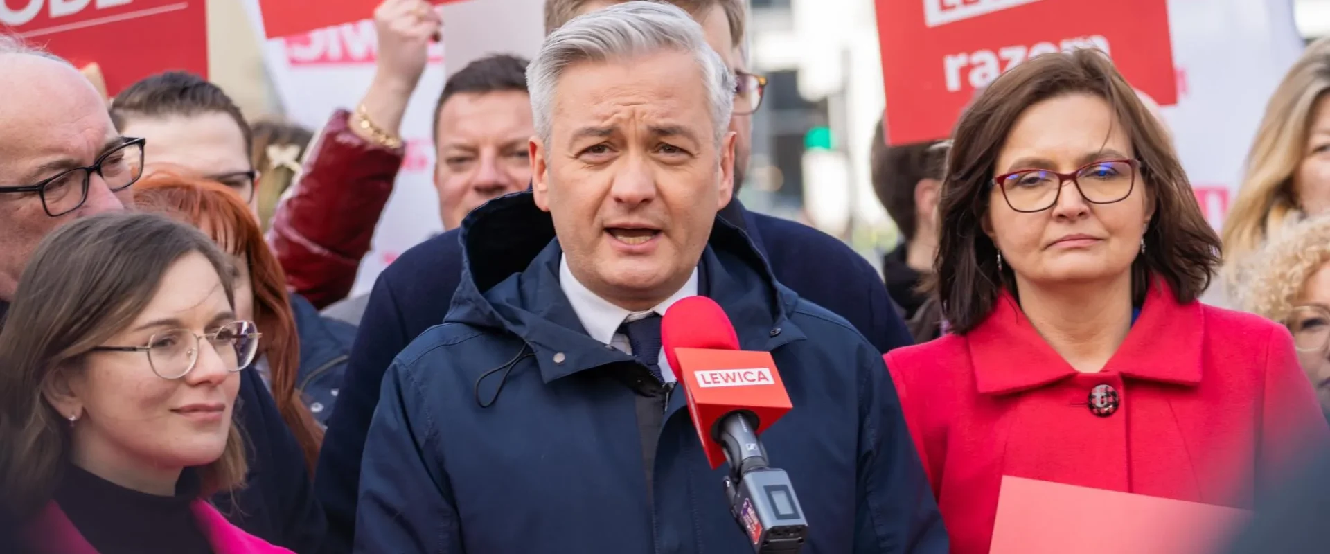 Mężczyzna w granatowej kurtce przemawia do mikrofonu podczas zgromadzenia, otoczony innymi uczestnikami i czerwonymi plakatami Lewica w tle.