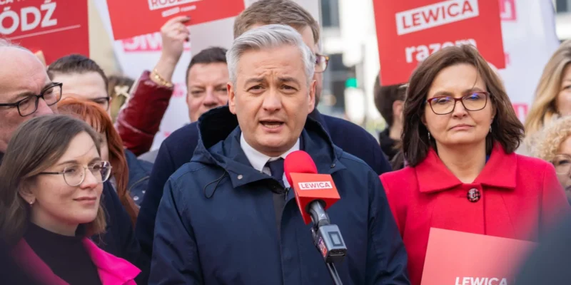 Mężczyzna w granatowej kurtce przemawia do mikrofonu podczas zgromadzenia, otoczony innymi uczestnikami i czerwonymi plakatami Lewica w tle.