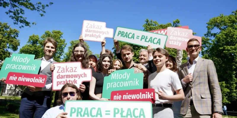 Grupa młodych ludzi stoi na zewnątrz i trzyma kolorowe plakaty z hasłami „PRACA = PŁACA” oraz „Zakaz darmowych praktyk”, protestując przeciw darmowym stażom.