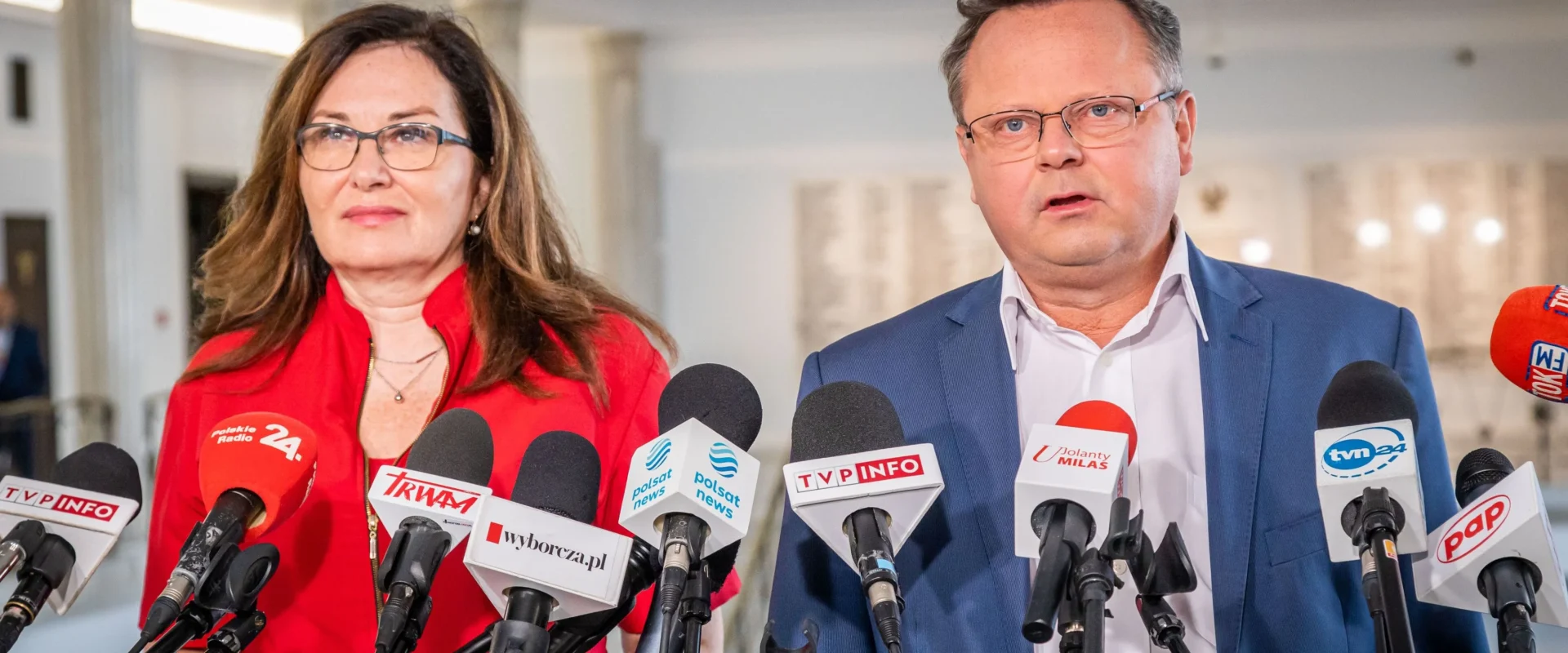 Konferencja prasowa w Pacanowie: para urzędników stoi przy licznych mikrofonach, kobieta w czerwonej bluzce i mężczyzna w granatowym garniturze.