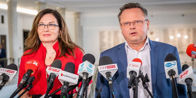 Konferencja prasowa w Pacanowie: para urzędników stoi przy licznych mikrofonach, kobieta w czerwonej bluzce i mężczyzna w granatowym garniturze.