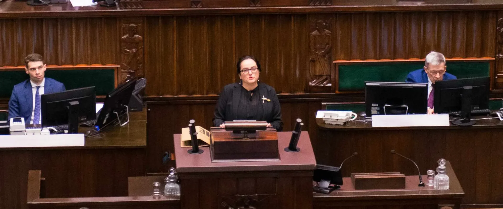 Kobieta stoi przy mównicy w sali plenarnej parlamentu, przemawiając do zgromadzonych przy biurkach z monitorami.
