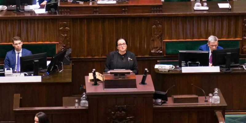 Kobieta stoi przy mównicy w sali plenarnej parlamentu, przemawiając do zgromadzonych przy biurkach z monitorami.