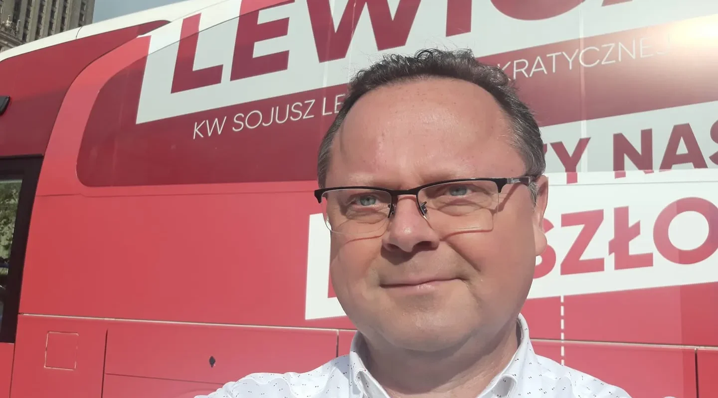 Mężczyzna w okularach robi selfie na tle dużego czerwonego autobusu z napisem Lewica.