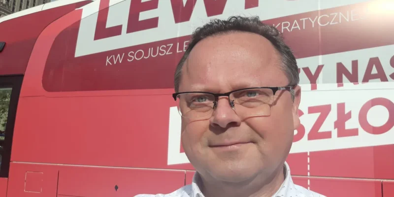 Mężczyzna w okularach robi selfie na tle dużego czerwonego autobusu z napisem Lewica.