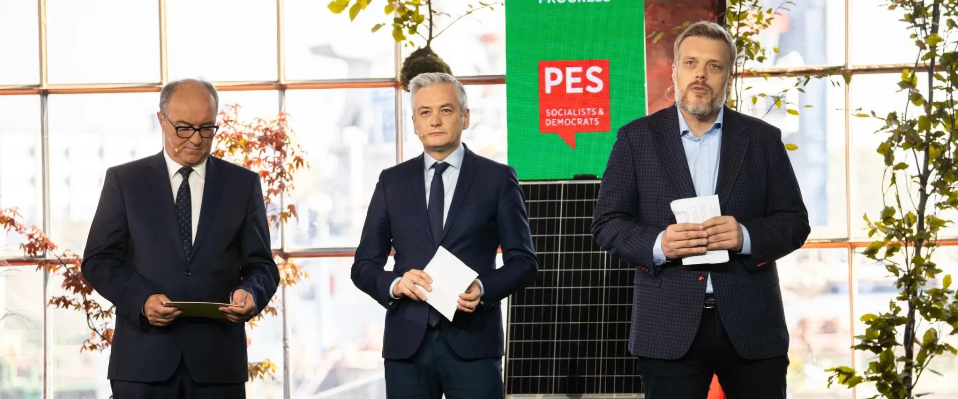 Na zdjęciu trzej mężczyźni stoją na scenie podczas wydarzenia, z zielonym tłem i roślinami w tle, prezentując dokumenty oraz podkreślając hasła związane z postępem i wartościami socjalistycznymi.