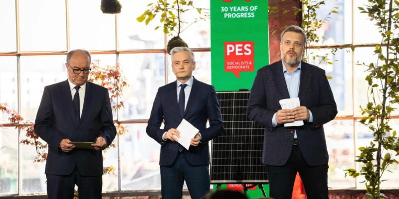 Na zdjęciu trzej mężczyźni stoją na scenie podczas wydarzenia, z zielonym tłem i roślinami w tle, prezentując dokumenty oraz podkreślając hasła związane z postępem i wartościami socjalistycznymi.