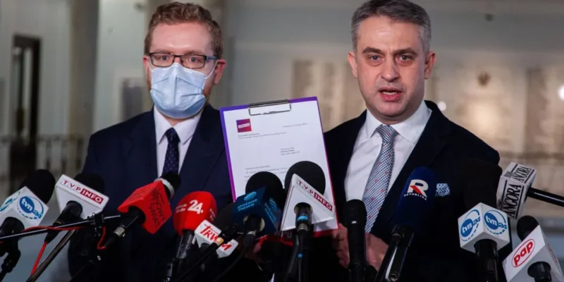 Dwaj mężczyźni w garniturach, jeden z maseczką, stoją przed mikrofonami i prezentują dokument podczas konferencji prasowej.