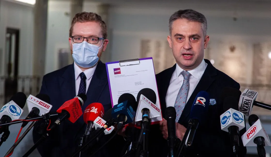 Dwaj mężczyźni w garniturach, jeden z maseczką, stoją przed mikrofonami i prezentują dokument podczas konferencji prasowej.