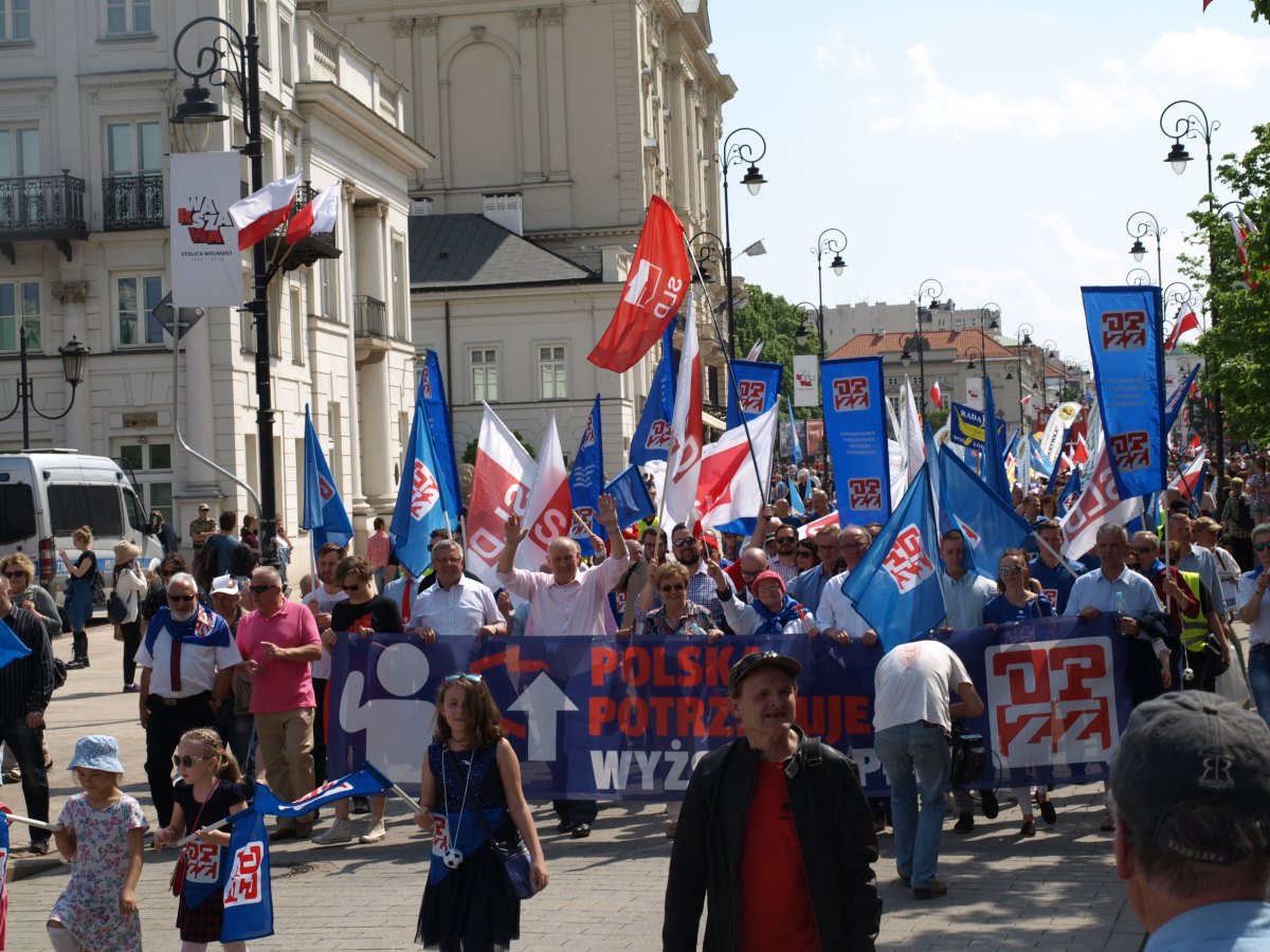 Tłum uczestników pochodu 1 Maja z flagami i banerami przechodzi ulicami Warszawy.