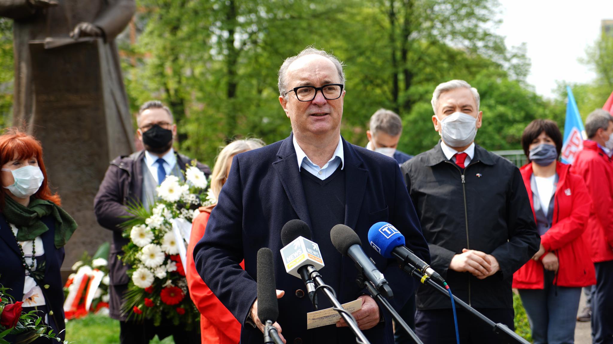 Na zdjęciu widoczny jest mężczyzna przemawiający do mikrofonów w towarzystwie innych osób, z kwiatami i pomnikiem w tle, podczas wydarzenia związanego z obchodami 1 Maja.