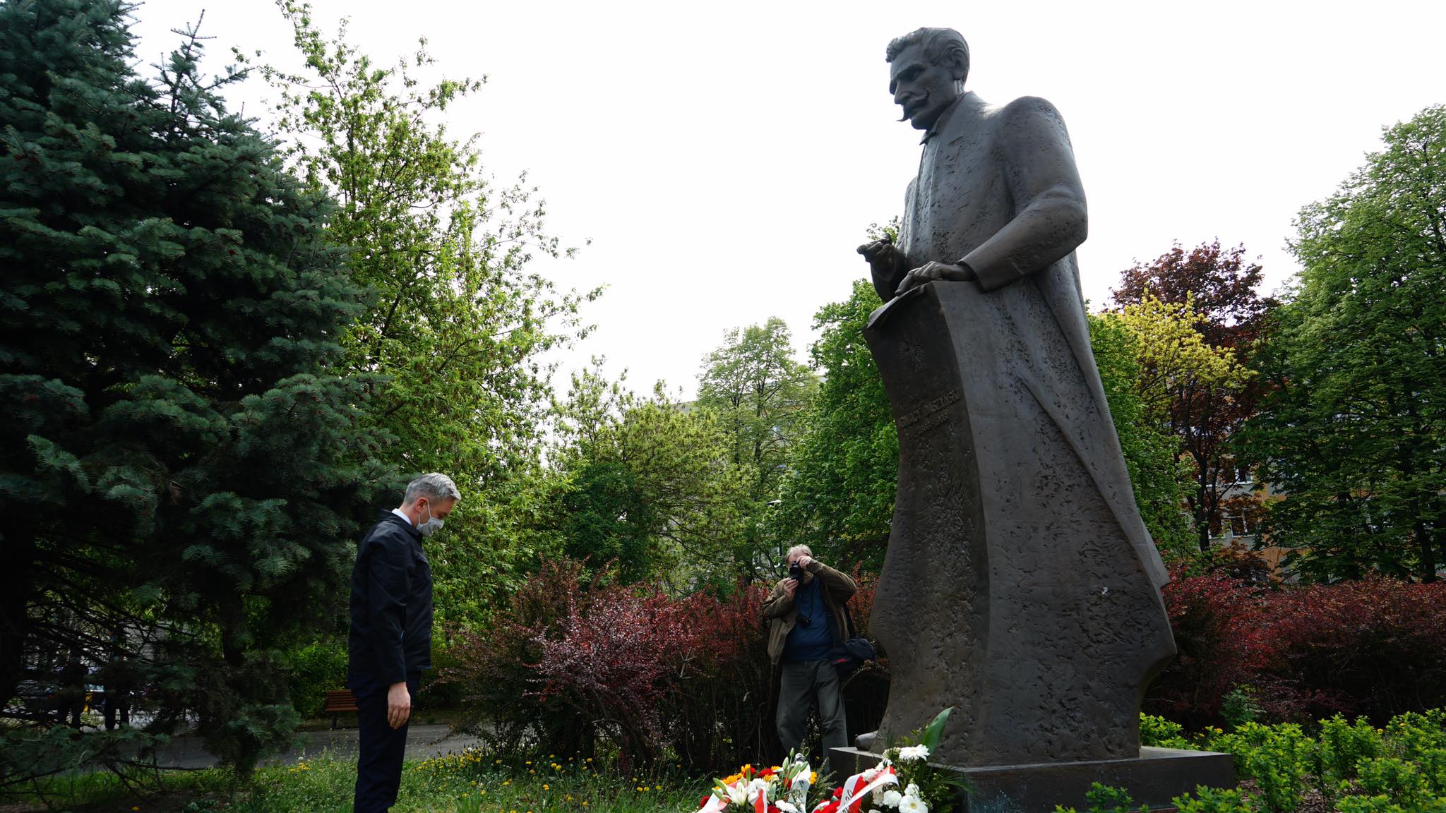Na zdjęciu widoczna jest postać mężczyzny składającego hołd przy pomniku, otoczonym zielenią i kwiatami.
