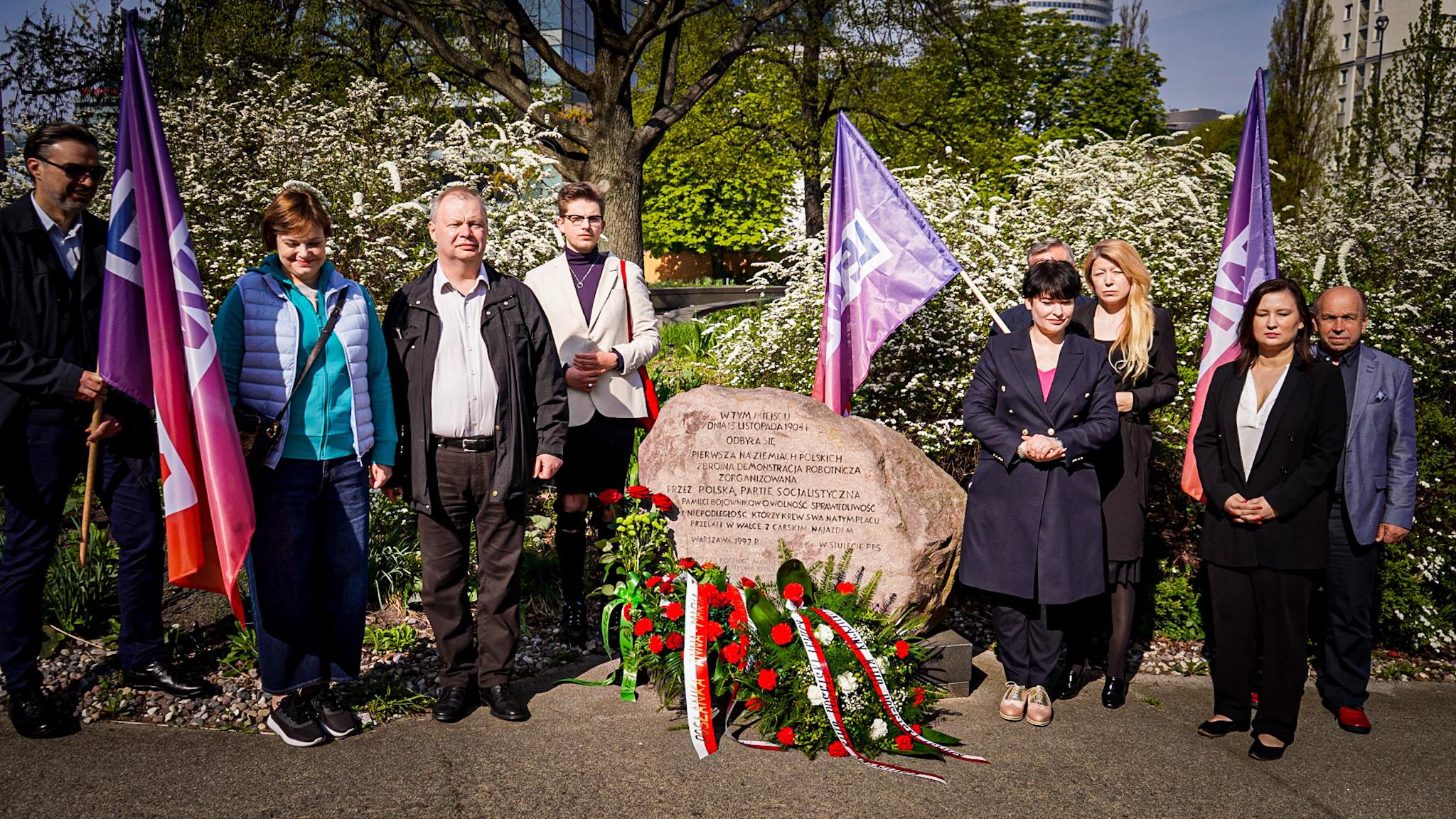 Grupa osób stoi w pobliżu kamienia pamiątkowego, na którym widnieje inskrypcja, otoczona kwiatami i flagami.
