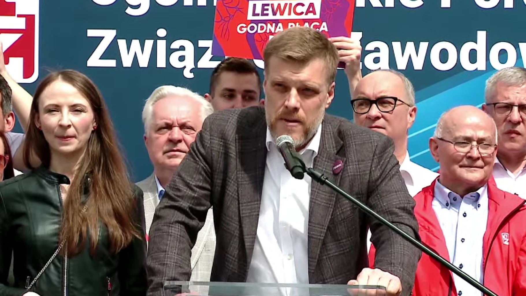 Na zdjęciu przedstawiony jest mężczyzna przemawiający z mównicy, otoczony przez grupę ludzi w tle, podczas wydarzenia związanego z Lewicą.