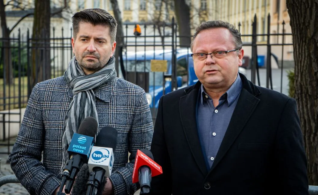 Dwóch mężczyzn stojących przy mikrofonach podczas konferencji prasowej na tle ogrodzenia, omawiających orzeczenie TSUE ws. pieniędzy za praworządność i ochrony funduszy unijnych przed rozkradaniem.