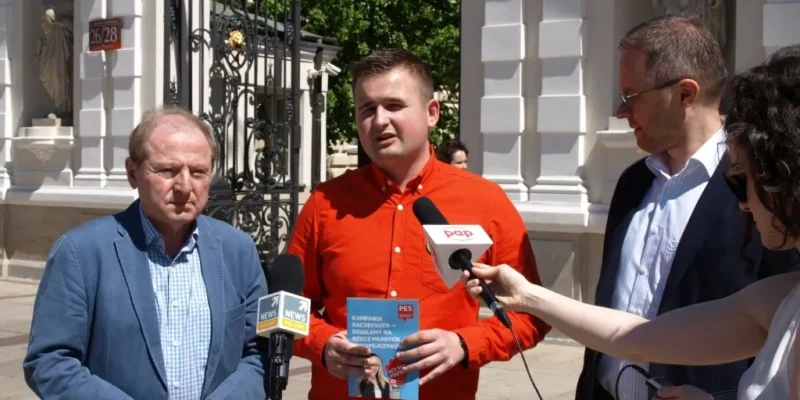 Przedstawiciele SLD podczas konferencji prasowej na zewnątrz — mężczyzna w czerwonej koszuli trzyma ulotkę i przemawia do mikrofonów.