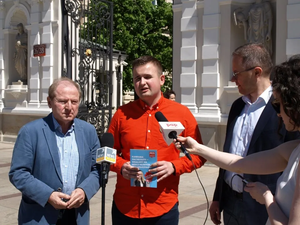 Przedstawiciele SLD podczas konferencji prasowej na zewnątrz — mężczyzna w czerwonej koszuli trzyma ulotkę i przemawia do mikrofonów.