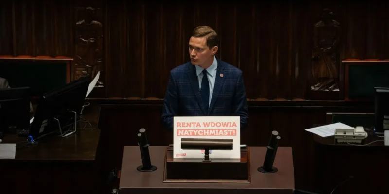 Polityk w garniturze stoi przy mównicy w sali plenarnej, prezentując plakat z napisem „Renta Wdowia Natychmiast!”.