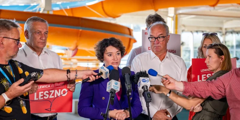 Kobieta w fioletowej marynarce przemawia do mikrofonów podczas konferencji prasowej, otoczona innymi politykami; w tle widoczne są banery Lewicy i pomarańczowe elementy konstrukcji.