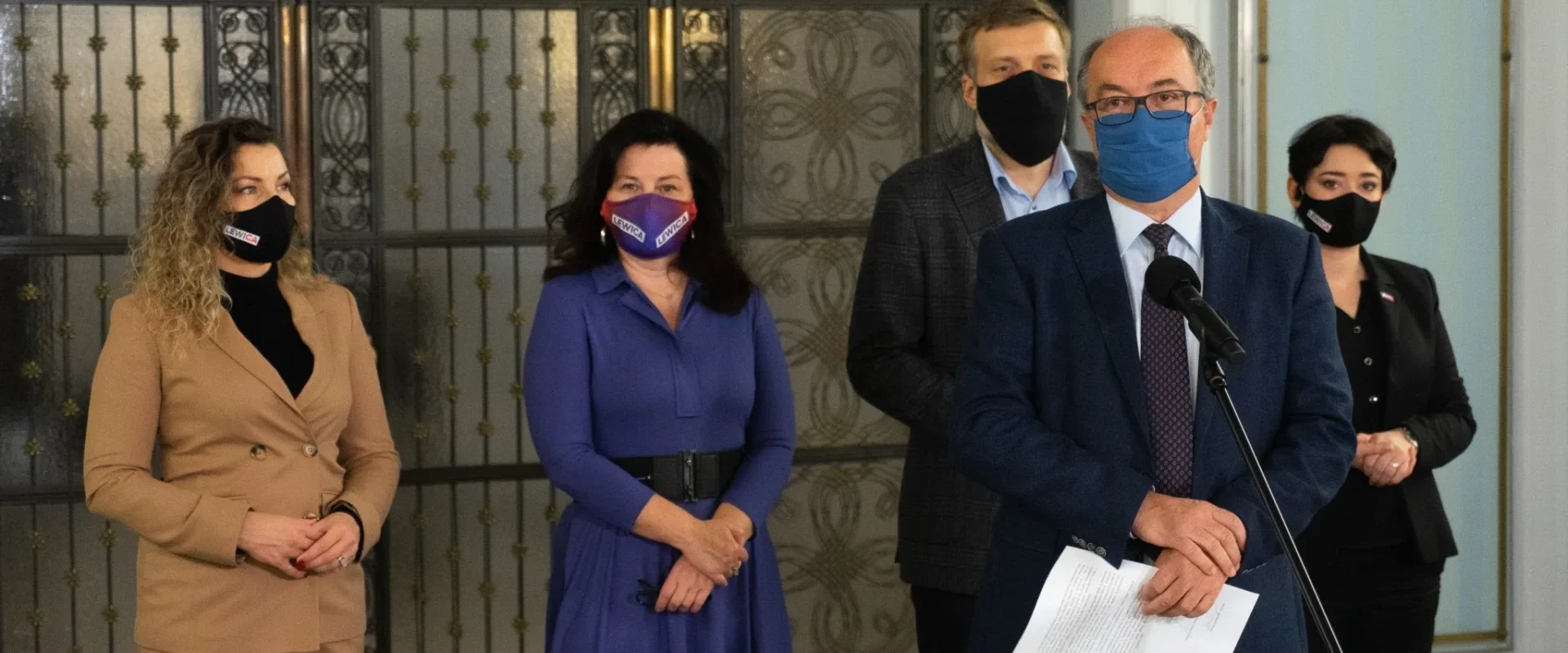 Grupa pięciu polityków Lewicy w maseczkach podczas konferencji prasowej, stojących przy mikrofonie w eleganckim wnętrzu z ozdobną metalową kratą w tle.