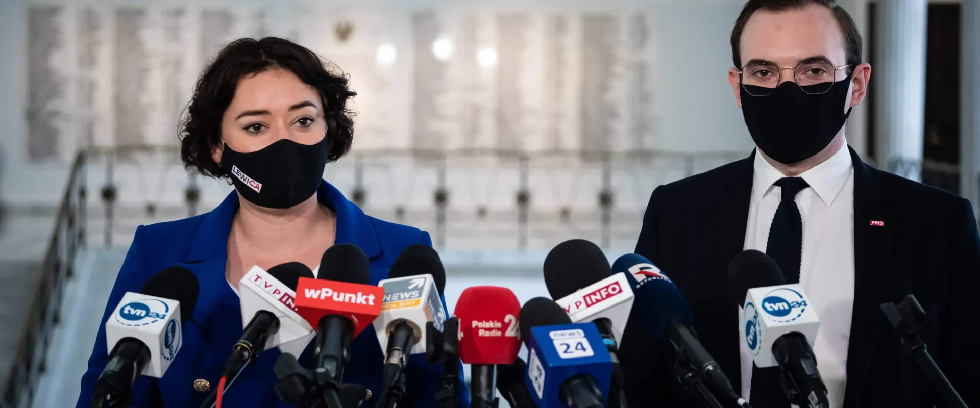 Konferencja prasowa dotycząca listu posłanki do ministra Czarnka: kobieta w niebieskim garniturze i mężczyzna w garniturze stoją za zestawem mikrofonów, oboje w maseczkach, w tle wnętrze budynku.
