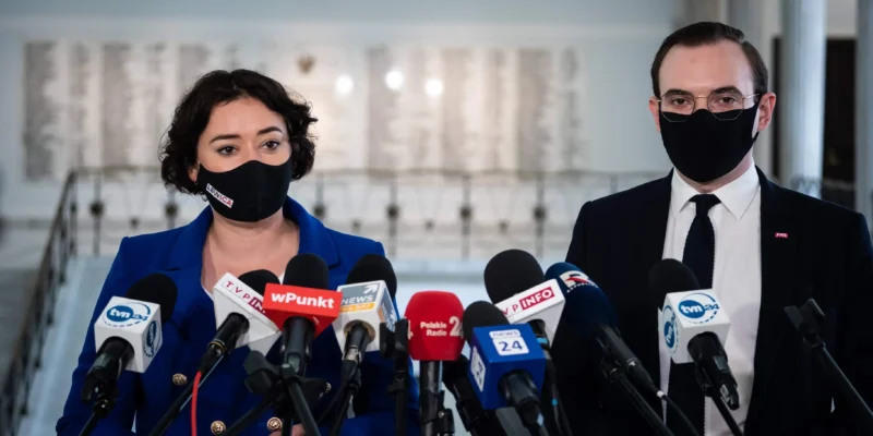 Konferencja prasowa dotycząca listu posłanki do ministra Czarnka: kobieta w niebieskim garniturze i mężczyzna w garniturze stoją za zestawem mikrofonów, oboje w maseczkach, w tle wnętrze budynku.