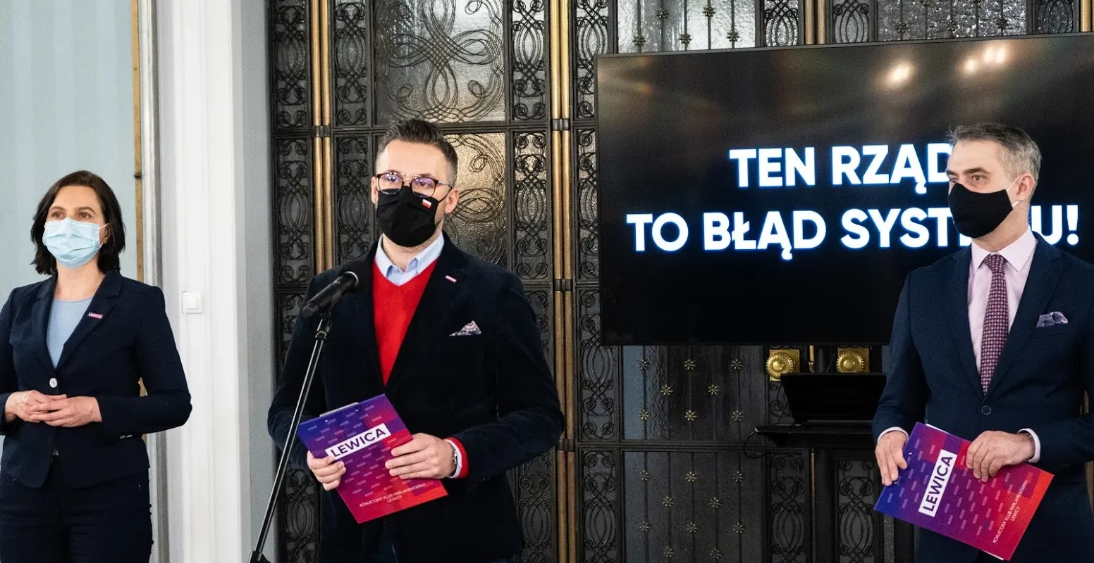 Trzy osoby w garniturach i maseczkach stoją przy mikrofonach podczas prezentacji, a na ekranie w tle widnieje hasło „TEN RZĄD TO BŁĄD SYSTEMU”.