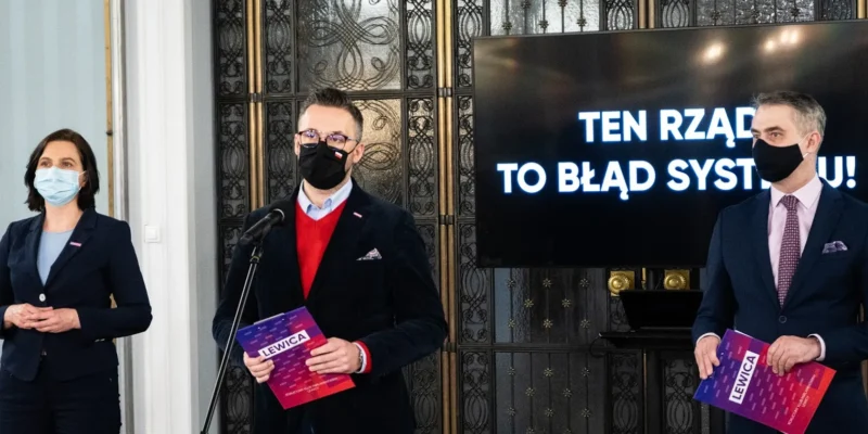 Trzy osoby w garniturach i maseczkach stoją przy mikrofonach podczas prezentacji, a na ekranie w tle widnieje hasło „TEN RZĄD TO BŁĄD SYSTEMU”.