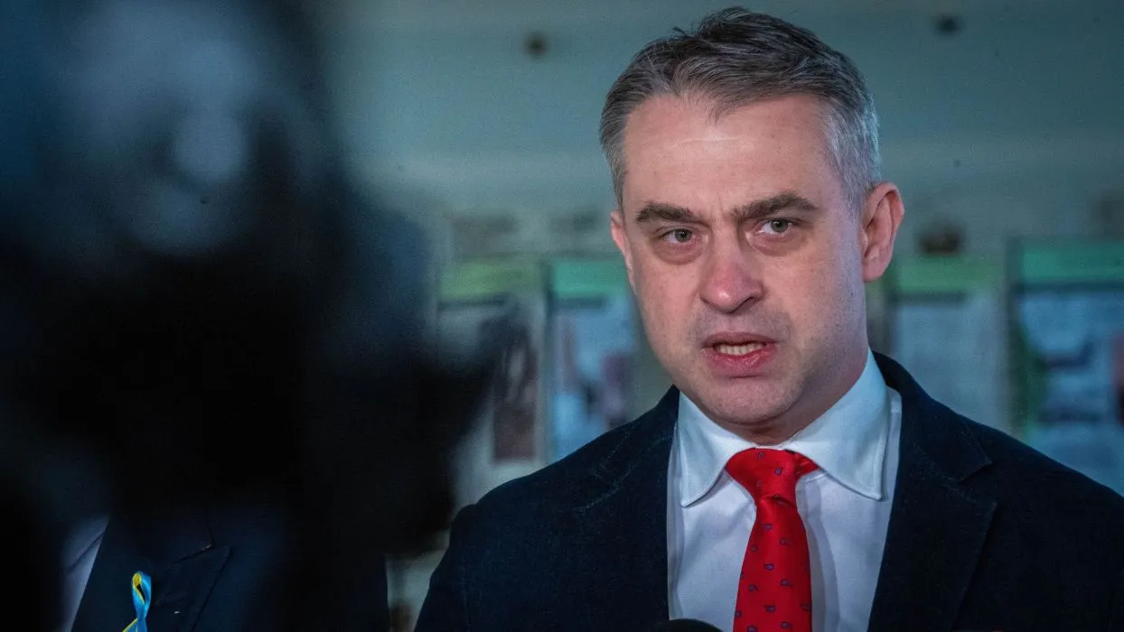 Mężczyzna w garniturze z czerwoną krawatem przemawia poważnie do mediów.