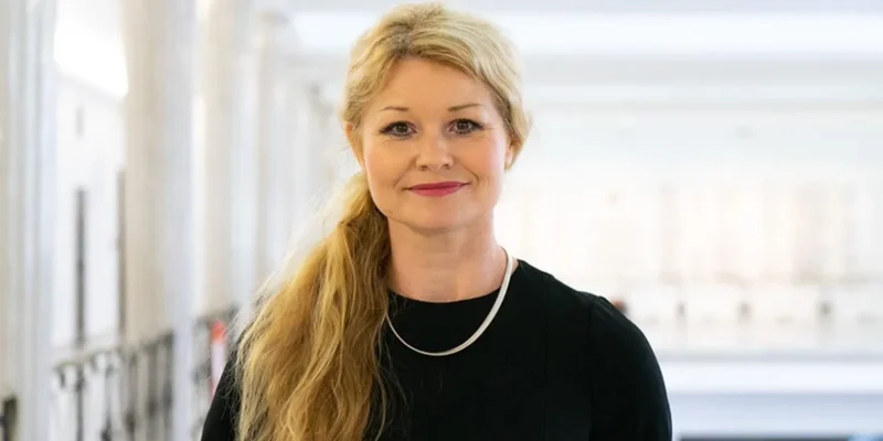 Kobieta z długimi, blond włosami uśmiecha się w eleganckim czarnym stroju, a w tle widać jasne wnętrze.