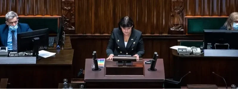 Na zdjęciu widać posiedzenie w parlamencie, z przewodniczącą siedzącą przy mównicy oraz dwoma mężczyznami w maseczkach, obserwującymi z miejsc.