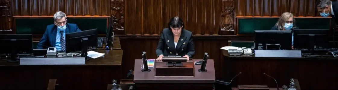 Na zdjęciu widać posiedzenie w parlamencie, z przewodniczącą siedzącą przy mównicy oraz dwoma mężczyznami w maseczkach, obserwującymi z miejsc.