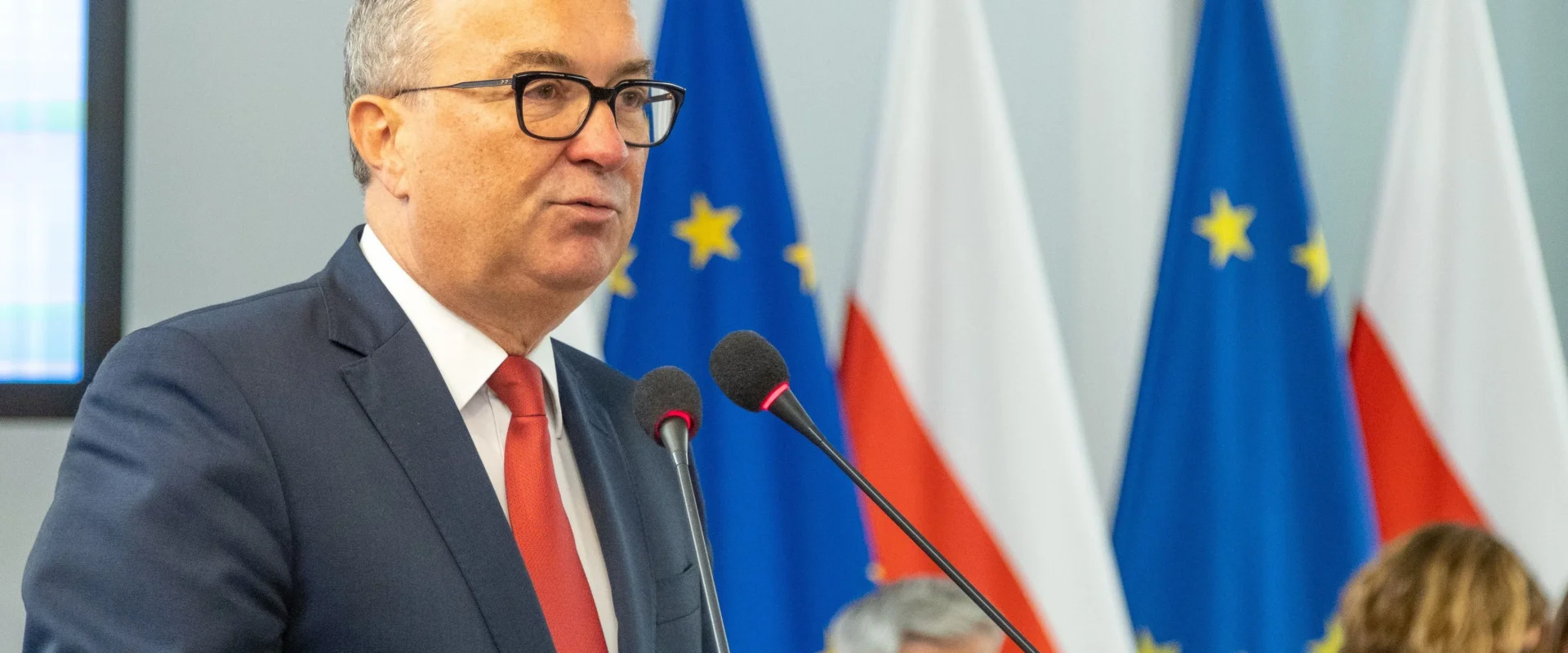 Mężczyzna w granatowym garniturze i czerwonym krawacie przemawia do mikrofonów podczas konferencji prasowej, z flagami Unii Europejskiej i Polski w tle.