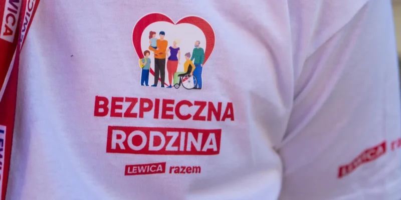 Na zdjęciu widoczna jest koszulka z napisem „BEZPIECZNA RODZINA” oraz grafiką przedstawiającą różnorodne postacie rodzinne, promującą inicjatywę „LEWICA razem”.