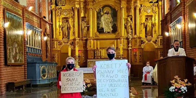 Dwie osoby stoją przed złoconym ołtarzem w kościele, trzymając duże plakaty z hasłami dotyczącymi praw kobiet, jedna w czerwonym płaszczu, druga w różowych spodniach, obie w maseczkach.