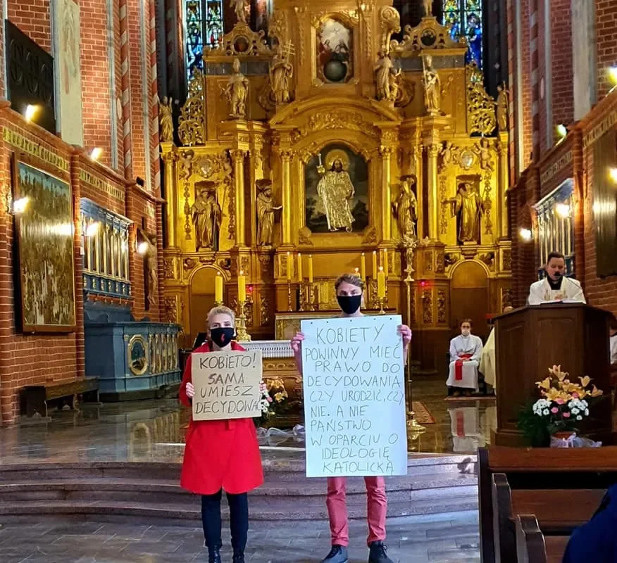 Dwie osoby stoją przed złoconym ołtarzem w kościele, trzymając duże plakaty z hasłami dotyczącymi praw kobiet, jedna w czerwonym płaszczu, druga w różowych spodniach, obie w maseczkach.