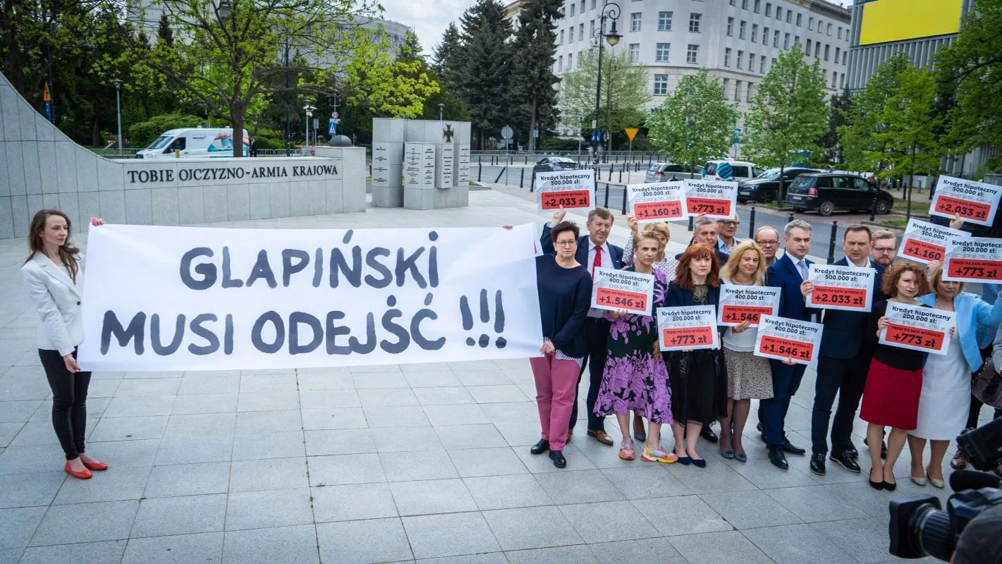 Grupa ludzi na placu trzyma duży baner z napisem „GLAPIŃSKI MUSI ODEJŚĆ!!!” oraz plakaty z kwotami kredytów hipotecznych.