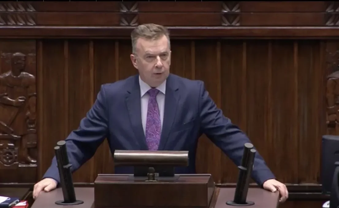 Mężczyzna w granatowym garniturze przemawia z mównicy w sali plenarnej.