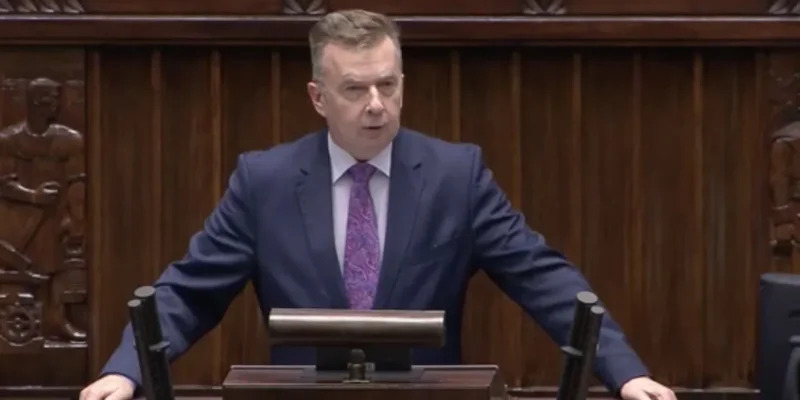 Mężczyzna w granatowym garniturze przemawia z mównicy w sali plenarnej.