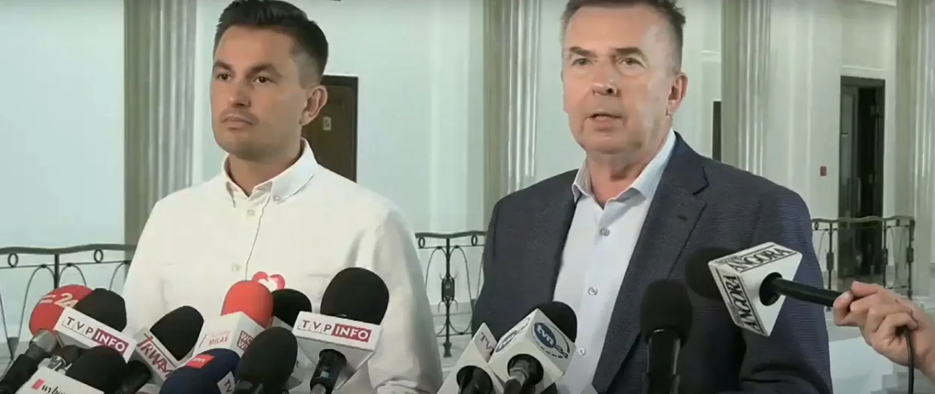 Dwóch mężczyzn, reprezentantów Lewicy i Koalicji Obywatelskiej, udziela konferencji prasowej, stojąc przy szeregu mikrofonów z logotypami stacji telewizyjnych w eleganckim wnętrzu.