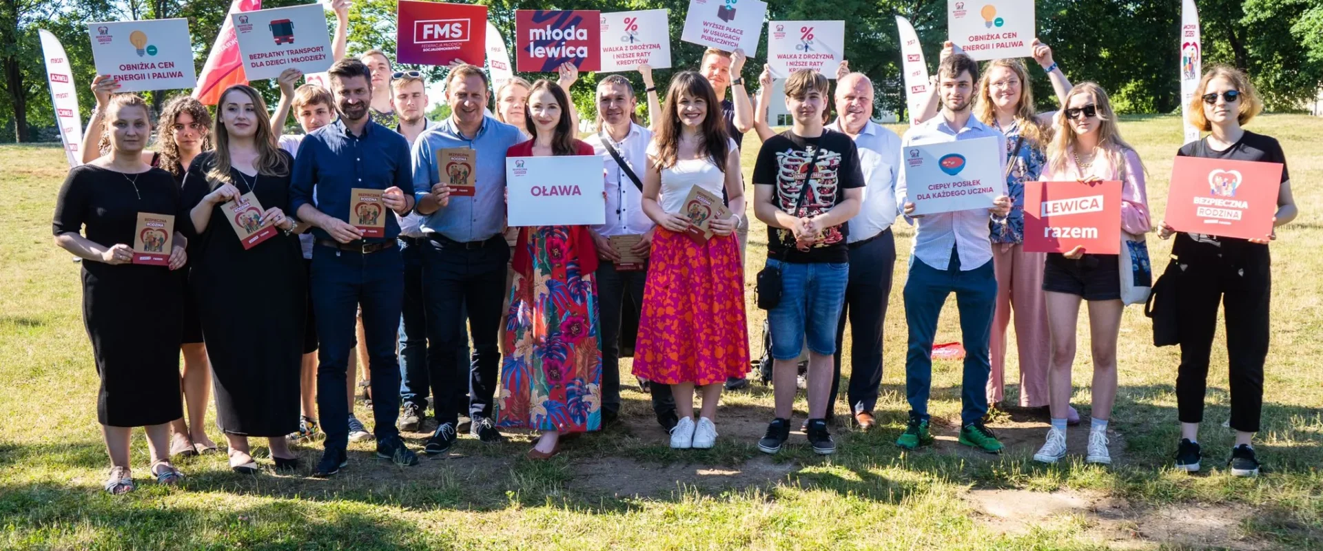 Grupa uczestników kampanii Lewicy stoi na zielonej trawie w parku, trzyma plakaty i materiały promocyjne z hasłami.