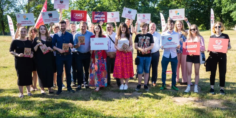 Grupa uczestników kampanii Lewicy stoi na zielonej trawie w parku, trzyma plakaty i materiały promocyjne z hasłami.